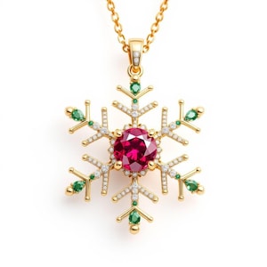 Könnte beinhalten: Goldfarbene Schneeflocken-Anhänger-Halskette mit einem großen, runden, roten Edelstein in der Mitte. Das Schneeflocken-Design hat kleine weiße und grüne Edelsteine und hängt an einer feinen Goldkette.