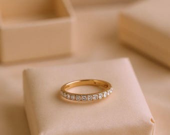 Può includere: Un anello a fascia in oro con una fila di piccoli diamanti a taglio quadrato. L'anello è appoggiato su una superficie di raso beige, con uno sfondo sfocato che include un portagioie. L'anello è un gioiello delicato.