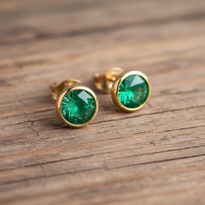 Könnte beinhalten: Ein Paar goldene Ohrstecker mit runden, smaragdgrünen Edelsteinen. Die Ohrringe sind in einer goldenen Lünettenfassung gefasst, und die Edelsteine haben ein facettiertes Aussehen. Die Ohrringe werden auf einer Holzoberfläche präsentiert.