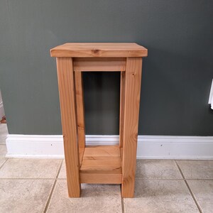 May include: A tall, rectangular wooden plant stand constructed from light-toned natural wood with a visible grain. The stand features a flat top surface, four sturdy vertical legs, and a lower shelf positioned between the legs near the base.