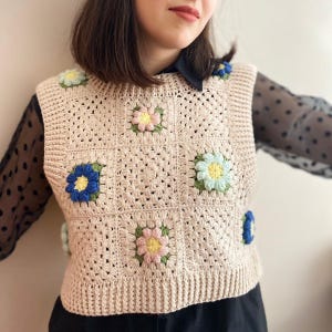 Può includere: Un gilet all'uncinetto beige ornato da applicazioni floreali colorate. I fiori presentano sfumature di blu, giallo, rosa e verde, aggiungendo un tocco giocoso al capo fatto a mano. Il gilet ha un motivo quadrato.