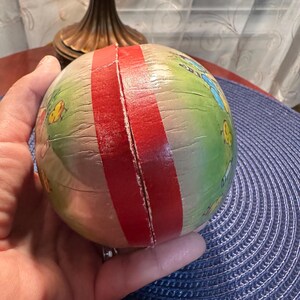 May include: A vintage, round paper toy with a red stripe and colorful illustrations of insects and flowers. The toy is held in a hand and has a slightly aged appearance.