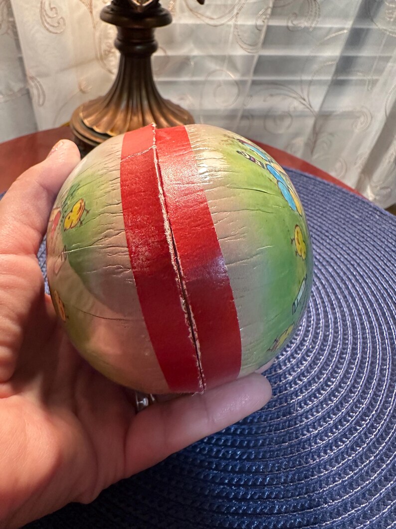 May include: A vintage, round paper toy with a red stripe and colorful illustrations of insects and flowers. The toy is held in a hand and has a slightly aged appearance.