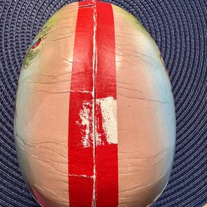 May include: An egg-shaped container with a red stripe down the center. The container has a light pink and beige color scheme with a cracked, aged appearance. The top of the container has a green and blue design.