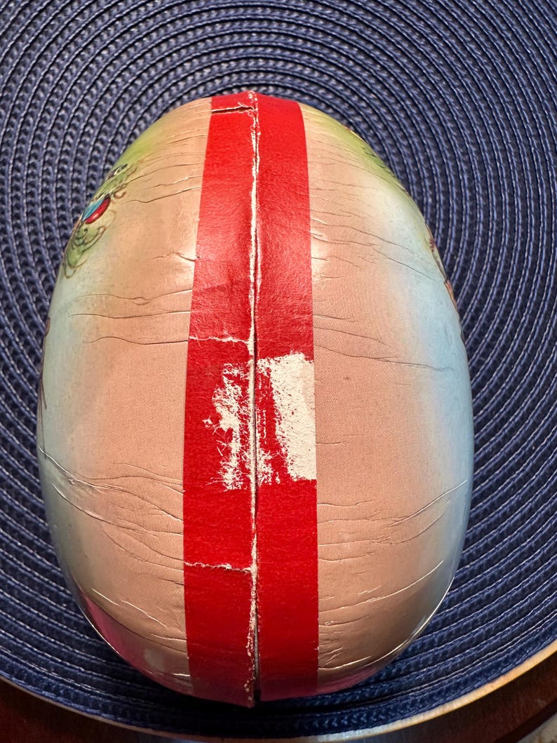 May include: An egg-shaped container with a red stripe down the center. The container has a light pink and beige color scheme with a cracked, aged appearance. The top of the container has a green and blue design.