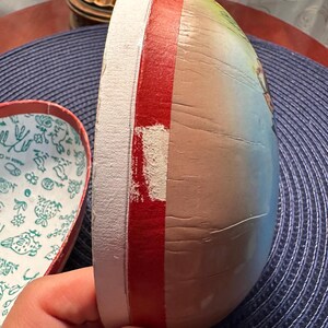 May include: A vintage, heart-shaped box with a lid. The box is painted with a colorful design and has a red stripe around the edge. The interior of the box is white with a floral pattern.