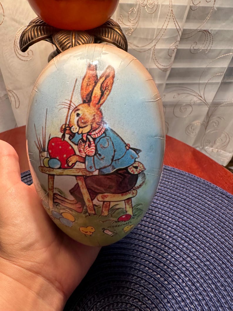 May include: An Easter egg with a painted illustration of a rabbit artist. The rabbit, wearing a blue jacket and brown pants, is painting a red egg. The egg has a light blue background with cracks.