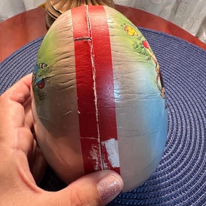May include: An egg-shaped decorative item with a red stripe down the center. The egg has a painted design with green, blue, and yellow hues. The item is held by a hand, and the background is a blue surface.