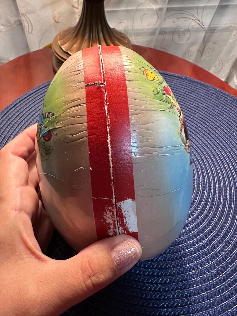 May include: An egg-shaped decorative item with a red stripe down the center. The egg has a painted design with green, blue, and yellow hues. The item is held by a hand, and the background is a blue surface.