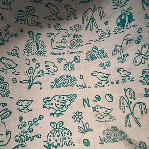 May include: Close-up of a white paper container with a teal Easter-themed pattern. The design includes bunnies, eggs, chicks, and the text "Made in Germany". The container has a slightly worn appearance.