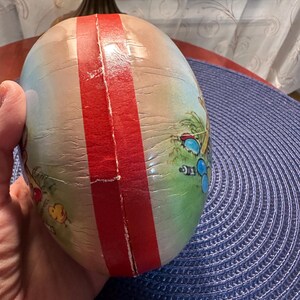 May include: An egg-shaped decorative item with a red stripe down the center. The egg features a painted scene with palm trees, beach umbrellas, and other beach-themed elements. The egg is held in a person's hand.
