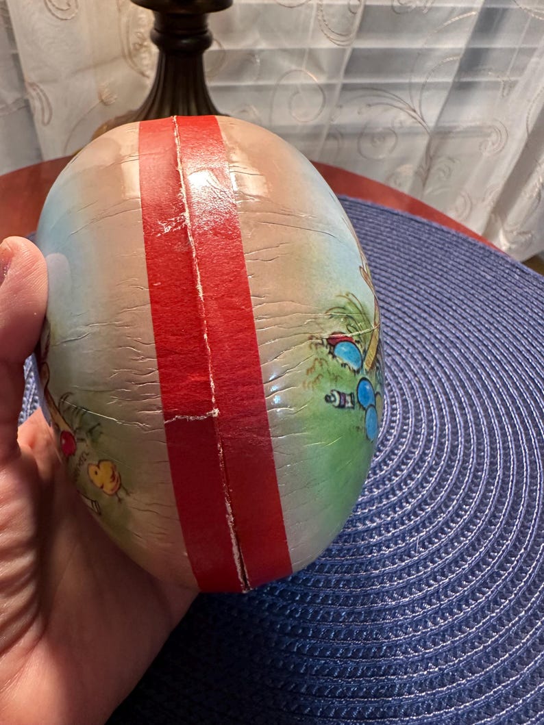 May include: An egg-shaped decorative item with a red stripe down the center. The egg features a painted scene with palm trees, beach umbrellas, and other beach-themed elements. The egg is held in a person's hand.