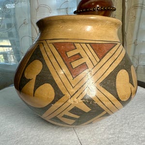 May include: A handcrafted, beige-colored pottery vase with a wide mouth and rounded body. The vase features geometric designs in black, red, and beige. The design includes angular shapes and curved elements. A small, decorative object sits on top.