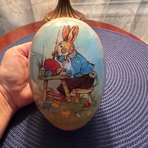 May include: An egg-shaped Easter decoration featuring a cartoon rabbit painting an Easter egg. The rabbit wears a blue jacket and sits at a small table. The egg has a light blue background with grass and other painted eggs.