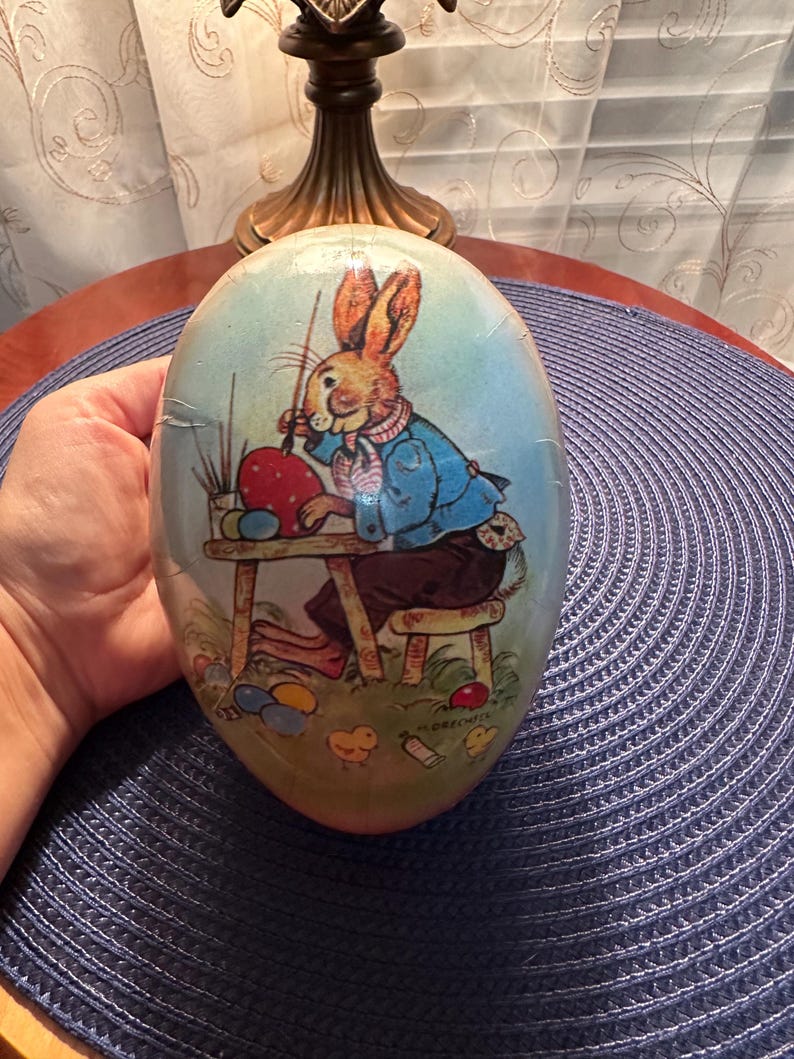 May include: An egg-shaped Easter decoration featuring a cartoon rabbit painting an Easter egg. The rabbit wears a blue jacket and sits at a small table. The egg has a light blue background with grass and other painted eggs.