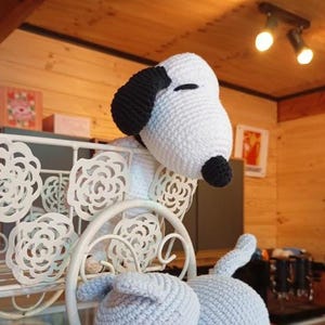 May include: A handmade crochet Snoopy plush toy, featuring a white head with black ears and nose, and a light blue body. The plush is positioned near a white decorative metal shelf with floral cutouts. The background includes wooden paneling and warm lighting.