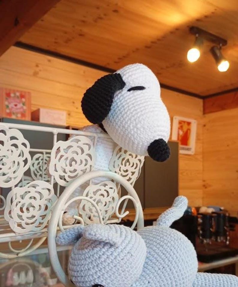Peut inclure: Une peluche Snoopy faite &agrave; la main au crochet, avec une t&ecirc;te blanche, des oreilles et un nez noirs, et un corps bleu clair. La peluche est plac&eacute;e pr&egrave;s d'une &eacute;tag&egrave;re d&eacute;corative en m&eacute;tal blanc avec des d&eacute;coupes florales. L'arri&egrave;re-plan comprend des boiseries et un &eacute;clairage chaleureux.