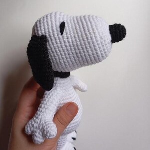Peut inclure: Une poup&eacute;e Snoopy au crochet, principalement blanche avec des d&eacute;tails noirs. Le chien a des oreilles noires, un nez noir et un collier noir. Les points de crochet sont visibles, donnant au jouet un aspect textur&eacute;. La poup&eacute;e est tenue dans une main.