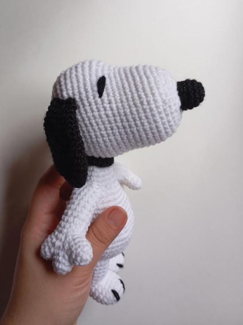 Peut inclure: Une poup&eacute;e Snoopy au crochet, principalement blanche avec des d&eacute;tails noirs. Le chien a des oreilles noires, un nez noir et un collier noir. Les points de crochet sont visibles, donnant au jouet un aspect textur&eacute;. La poup&eacute;e est tenue dans une main.