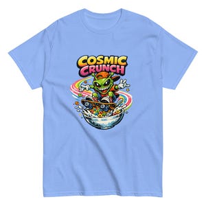Könnte beinhalten: Hellblaues T-Shirt mit einem Cartoon-Alien, das auf einer Schüssel Müsli skateboardet. Das Alien ist grün mit einem roten Hut und fährt auf einem Regenbogen Skateboard. Der Text "COSMIC CRUNCH" steht in Gelb und Pink über dem Alien.