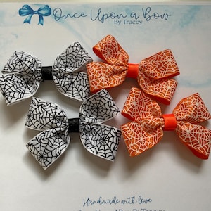 May include: Six handmade Halloween hair bows. Three bows feature white fabric with black spiderwebs, and three are orange with white spiderwebs. The bows are on a white background with the text "Once Upon a Bow by Tracey".