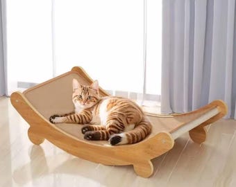 Könnte beinhalten: Eine getigerte Katze liegt auf einer Katzenhängematte aus Holz. Die Hängematte hat ein beiges Stoffbett und einen geschwungenen Rahmen aus Naturholz. Die Katze hat braune und weiße Streifen. Die Hängematte ist für den Innenbereich konzipiert.