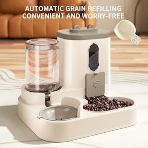 Puede incluir: Un comedero automático para mascotas en crema y gris, con el texto "AUTOMATIC GRAIN REFILLING CONVENIENT AND WORRY-FREE". Cuenta con un depósito de agua transparente, un dispensador de comida y un cuenco. El dispensador está lleno de pienso marrón.