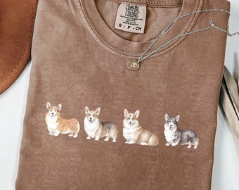 May include: Espresso-colored t-shirt with a crew neck and a graphic of four corgi dogs. The shirt has a Comfort Colors tag and a silver necklace with a pendant. The shirt is on a white surface.