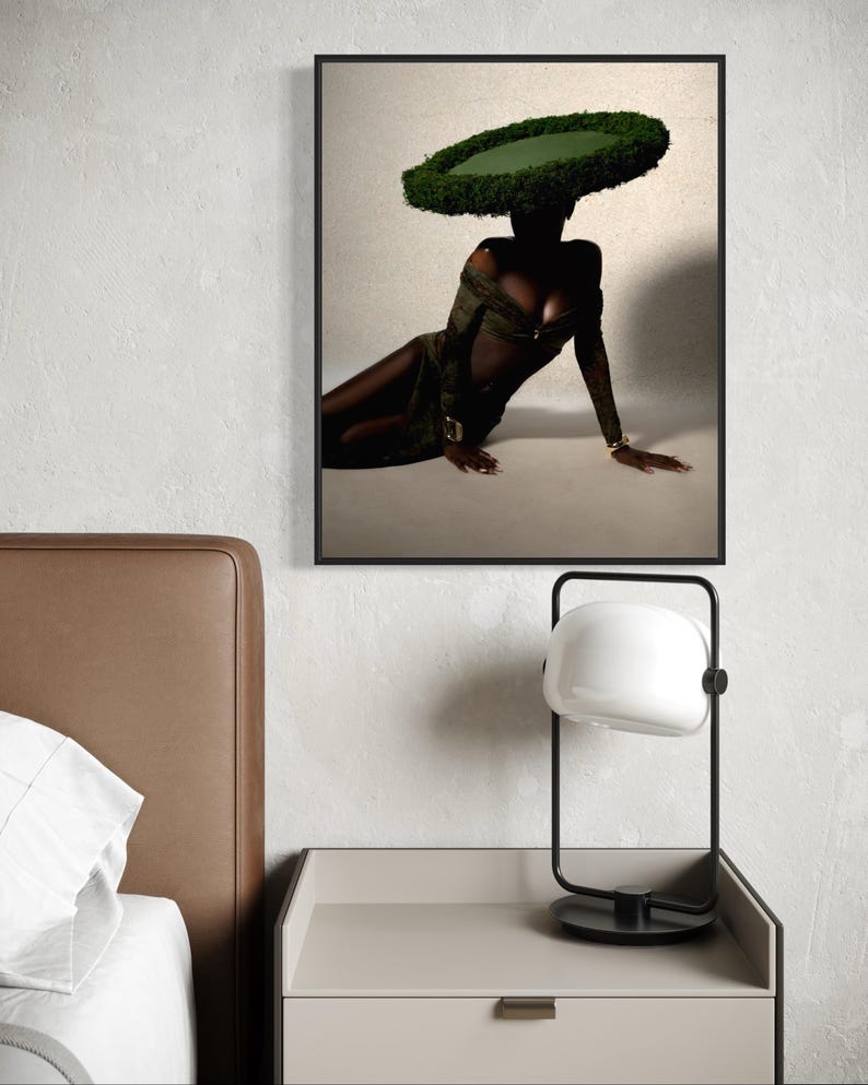 May include: A framed art print featuring a person wearing a large, circular green hat and a dark green dress. The person is seated on a light background. A black and white lamp sits on a nightstand.