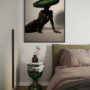 May include: A framed art print featuring a person wearing a green dress and a large, circular, green hat. The person is reclining on a light-colored surface. A green, hourglass-shaped side table holds books and a vase with flowers.