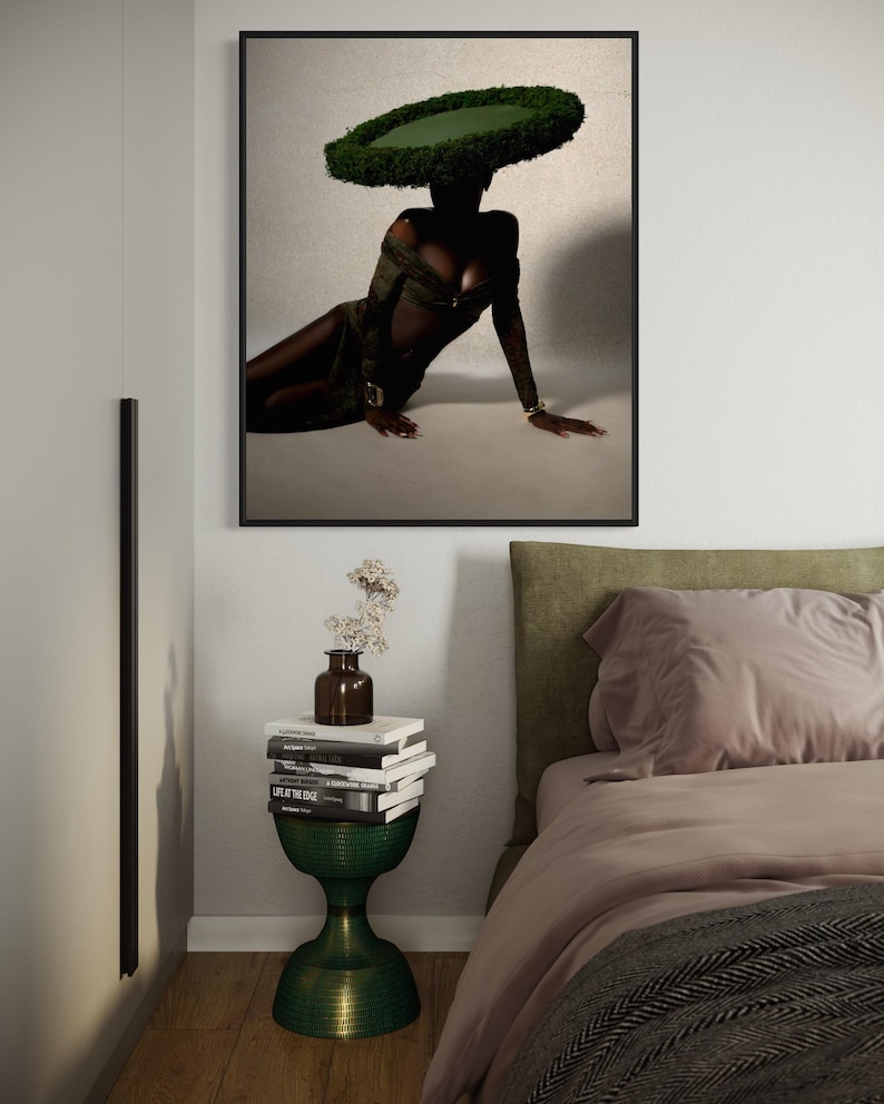 May include: A framed art print featuring a person wearing a green dress and a large, circular, green hat. The person is reclining on a light-colored surface. A green, hourglass-shaped side table holds books and a vase with flowers.