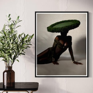 May include: A framed art print featuring a person wearing a large, circular green headpiece. The person is dressed in a dark green outfit and is seated. The artwork is displayed on a white wall next to a vase with green leaves.