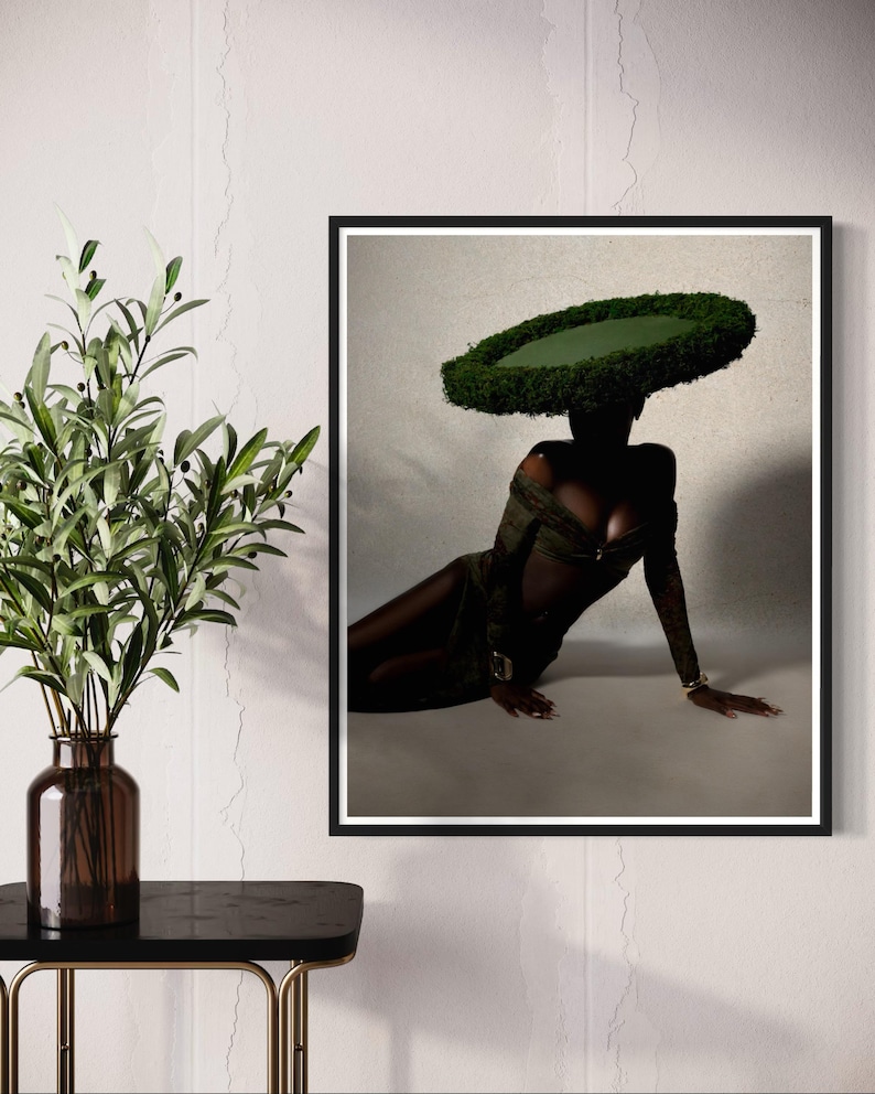 May include: A framed art print featuring a person wearing a large, circular green headpiece. The person is dressed in a dark green outfit and is seated. The artwork is displayed on a white wall next to a vase with green leaves.