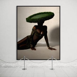 May include: A framed art print featuring a person wearing a green dress and a large, circular green hat. The person is reclining on a light-colored surface. The artwork is displayed in a gallery setting.