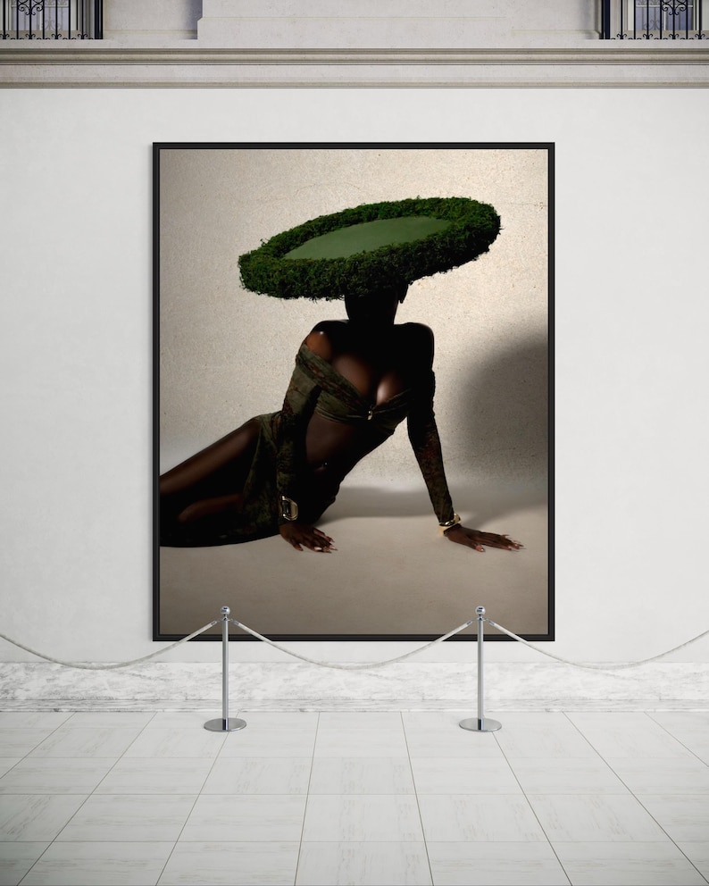 May include: A framed art print featuring a person wearing a green dress and a large, circular green hat. The person is reclining on a light-colored surface. The artwork is displayed in a gallery setting.