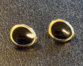 May include: A pair of black and gold earrings. Each earring features a glossy, oval-shaped black stone set within a gold-colored frame. The earrings are displayed on a textured, dark surface.