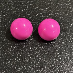 May include: Two bright pink, spherical objects are displayed against a dark, textured background. The spheres have a smooth, glossy surface, reflecting light. The image is a close-up, highlighting the color and shape of the objects.