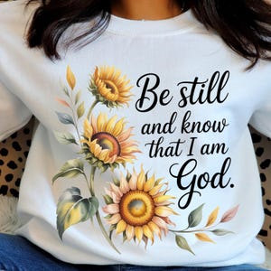 Peut inclure: Sweat-shirt blanc avec un motif aquarelle de tournesols et le texte "Be still and know that I am God." Les tournesols sont jaunes, oranges et bruns avec des feuilles vertes. Le texte est en écriture noire.