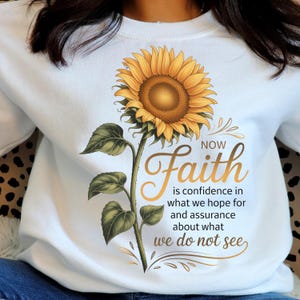 Peut inclure: Sweat-shirt blanc avec un motif de tournesol, feuilles et tige vertes. Le texte doré dit "Faith is confidence in what we hope for and assurance about what we do not see".