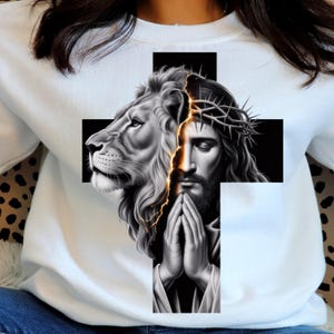 Peut inclure: Sweat-shirt blanc avec un motif graphique. Le motif représente une tête de lion et une représentation de Jésus portant une couronne d'épines, sur une croix noire. L'œuvre est en niveaux de gris avec un effet d'éclair doré.