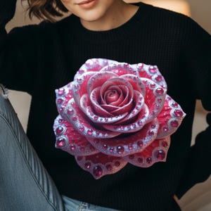 Peut inclure: Pull-over noir avec une grande rose ornementale. La rose est rouge vif avec des détails argentés et des pierres roses incrustées. Un vêtement élégant et accrocheur.