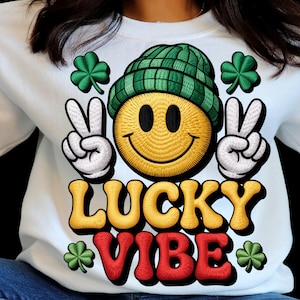 May include: White sweatshirt with a large embroidered design featuring a yellow smiley face wearing a green beanie and making peace signs. The words "LUCKY VIBE" are in yellow and red, with green shamrocks.