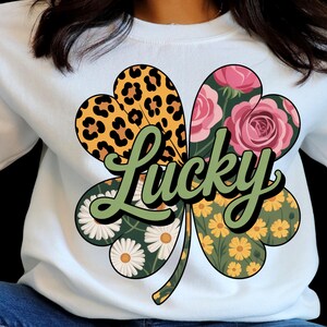 Peut inclure: Sweat-shirt blanc avec un motif de trèfle à quatre feuilles. Les feuilles du trèfle présentent un imprimé léopard, des roses roses, des marguerites et des fleurs jaunes. Le mot "Lucky" est écrit en vert au centre.