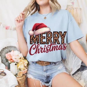 Peut inclure: T-shirt bleu clair avec l'inscription "MERRY Christmas" dans un design festif. Le mot "MERRY" présente des lettres à imprimé léopard, et un bonnet de Père Noël est au-dessus. Le mot "Christmas" est en écriture rouge.