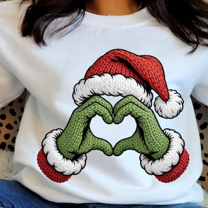 Peut inclure: Sweat-shirt blanc avec un motif de bonnet de Père Noël rouge et blanc au-dessus de mains vertes formant un cœur. Les mains ont une texture tricotée verte et des poignets rouges. Un design de vacances festif.