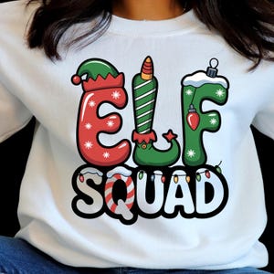 May include: White sweatshirt with the words "ELF SQUAD" in a festive design. The letters of "ELF" are decorated like an elf hat, a candy cane, and a Christmas ornament. The word "SQUAD" is in a candy cane font.