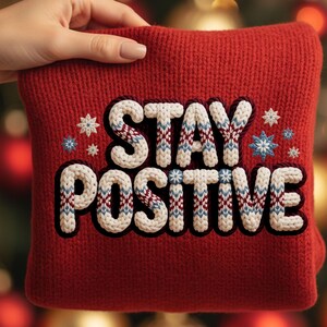 May include: A red knitted cushion with the words "STAY POSITIVE" embroidered in white, blue, and burgundy yarn. The cushion also features embroidered snowflake designs. The cushion is held by a hand.