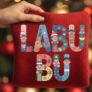 May include: A red knitted sweater with the word "LABU BU" in colorful, illustrated letters. Each letter features a unique design with cartoon characters and holiday-themed elements like trees and snowflakes. The sweater is held by a hand.