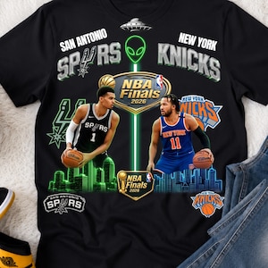 Peut inclure: Un t-shirt noir avec un graphisme pour les finales NBA 2026 entre les San Antonio Spurs et les New York Knicks. Le design comprend des images de deux joueurs de basket, des logos d'équipe, une icône extraterrestre et le texte NBA Finals 2026.