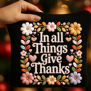 May include: Black cushion with floral embroidery and the text "In all Things Give Thanks." The embroidery features white, pink, yellow, and purple flowers, green leaves, and small pink hearts. The cushion is held by a hand.